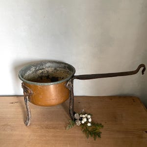 Peut inclure: Une casserole ancienne en cuivre avec une longue poignée incurvée et des pieds décoratifs. La casserole présente une patine ternie, montrant des signes d'âge et d'utilisation. Un petit arrangement de verdure et de baies blanches repose près de la base.