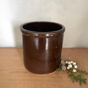 May include: A cylindrical stoneware crock featuring a deep, glossy dark brown glaze. The vessel has a thick, rounded rim and a smooth, uniform exterior surface. The pot is open at the top, revealing a hollow interior, and sits upright.