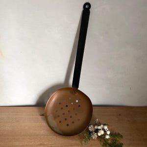 Puede incluir: Un espumadera de color cobre con un mango negro. El cabezal redondo tiene varios agujeros pequeños. El mango tiene un lazo para colgar. El utensilio de cocina está diseñado para colar alimentos de líquidos. Mide unos 30 cm de largo.