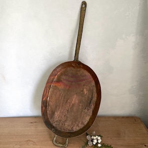 May include: An antique copper pan with a long, brass handle and a smaller handle on the base. The pan has a rounded, oval shape and shows signs of age and use. The copper has a rich, warm colour with areas of discolouration.