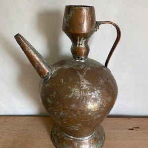 May include: An antique copper pitcher with a rounded body, a long spout, and a handle. The pitcher has a worn, aged patina and intricate etched designs.