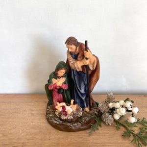 May include: A Nativity scene figurine depicting Mary, Joseph, and baby Jesus in a manger. Mary is kneeling in a red and green robe, Joseph stands with a staff, and baby Jesus is in a woven basket. The figures are on a brown base.