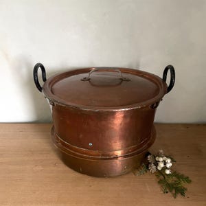 May include: A vintage copper double boiler with black handles. The boiler has a round shape and is made of two pieces, a pot and a lid. The pot is sitting on a wooden surface.