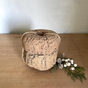May include: A cylindrical, light brown, textured cork candle holder with a central opening. The cork is wrapped with thin, natural-colored twine. A small decorative sprig of green pine needles and white berries is placed next to the base.