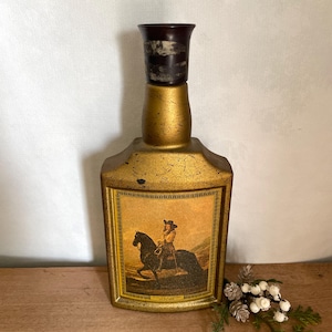 May include: Vintage gold-colored bottle with a dark brown stopper. The bottle features a framed image of a person on horseback. The bottle has a textured, aged appearance, suggesting it is an antique or collectible item.