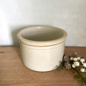 Puede incluir: Un recipiente de cerámica cilíndrico con un esmalte blanquecino moteado y un borde redondeado suave. La pieza presenta una sutil hendidura horizontal cerca del borde superior, mostrando un diseño simple y funcional con una paleta de colores neutros.