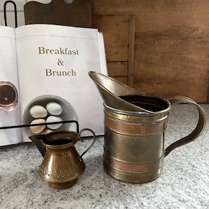 May include: Two brass pitchers, one small and one large, with copper accents. The larger pitcher has a handle and a spout. The smaller pitcher has a handle and a decorative design.