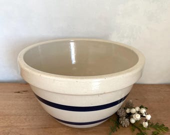 Puede incluir: Un cuenco mezclador de gres color crema con un borde ancho y dos rayas azul marino. El cuenco está sobre una superficie de madera, con una ramita de vegetación y bayas blancas. Un artículo de cocina clásico.
