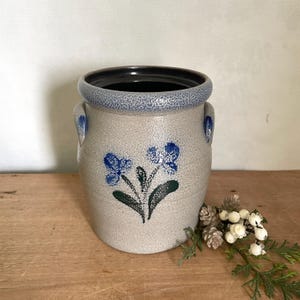 May include: A cylindrical stoneware crock with a speckled light gray body, featuring a thick blue speckled rim and two small side handles. The front is decorated with a hand-painted blue floral design with dark green leaves on the center surface.