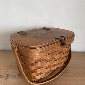 May include: A brown wooden basket with a lid and a woven handle. The lid has a dark brown leather strap and a small metal latch. The basket has a woven design and is made of natural wood.