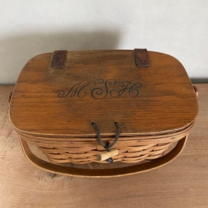 May include: A wooden basket with a brown lid and a woven base. The lid has a leather strap and a black leather handle. The basket has a black leather handle and a wooden knob. The lid has the initials "M.S.H." engraved on it.