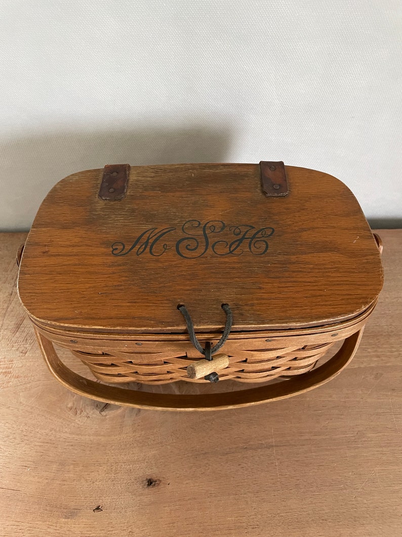 May include: A wooden basket with a brown lid and a woven base. The lid has a leather strap and a black leather handle. The basket has a black leather handle and a wooden knob. The lid has the initials "M.S.H." engraved on it.