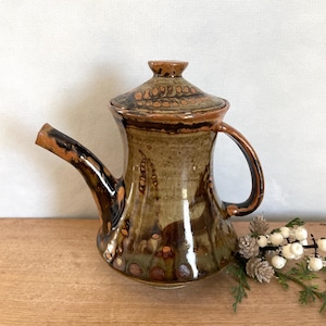 May include: A handcrafted ceramic teapot with a lid. The teapot features a brown and green mottled glaze with a dark brown handle and spout. The lid has a decorative pattern. The teapot is on a wooden surface.