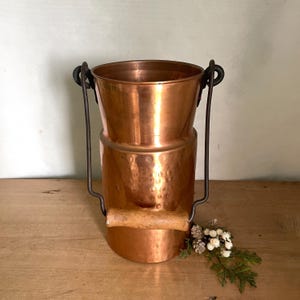 May include: A cylindrical copper vessel with a hammered texture on the lower half and a smooth upper section. It features a dark metal swing handle with a light-colored wooden grip attached to two circular loops on the rim of the container.