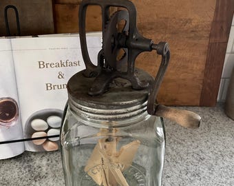 Könnte beinhalten: Antike Butterquirn mit einem Glasgefäß und einem Holzgriff. Das Gefäß trägt ein Etikett mit der Aufschrift "CHURN & SWIRL".