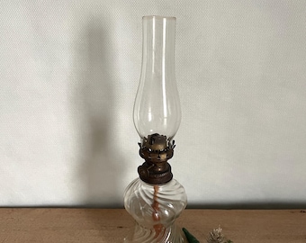 May include: A clear glass oil lamp with a twisted base and a brass wick holder. The lamp is sitting on a wooden surface.