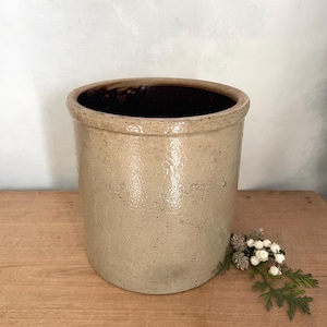 May include: A cylindrical stoneware crock featuring a speckled beige exterior finish and a smooth, dark brown glazed interior. The vessel has a thick, rounded rim and a sturdy base, presenting a neutral, earthy appearance with a textured surface.