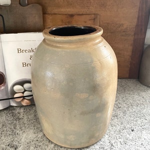 May include: A large, grey stoneware crock with a wide mouth and a slightly rounded body. The crock has a dark brown glaze on the inside rim and a light brown glaze on the outside.