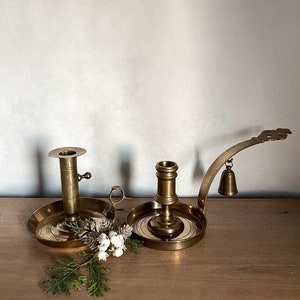 May include: Two antique brass candle holders. One is a simple candlestick with a round base and a single candle holder. The other is a more elaborate candlestick with a curved arm and a bell hanging from it.