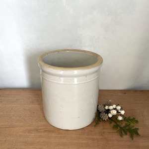 Peut inclure: Un pot en grès cylindrique de couleur blanc cassé avec une finition émaillée lisse et un bord épais non émaillé. Le récipient présente un design simple à parois droites avec des variations de surface subtiles et un ton neutre.
