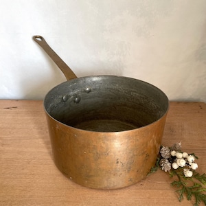 Könnte beinhalten: Ein Vintage-Kochtopf aus Kupfer mit einem langen Messinggriff. Der Topf hat einen breiten, abgerundeten Körper mit einer angelaufenen Patina, die Gebrauchsspuren aufweist. Der Innenraum ist aus dunklerem, kontrastierendem Metall. Ein dekorativer Zweig mit Grün und Beeren liegt in der Nähe.