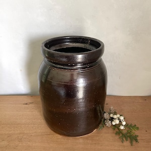 Op de afbeelding: Een donkerbruine, cilindrische keramische pot met een dikke, afgeronde rand en een gladde, glanzende afwerking. Het vat heeft een brede opening en een stevig, rechtwandig lichaam met een diepe, uniforme kleur over het gehele oppervlak.
