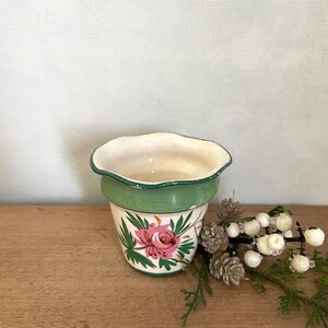 May include: A small, ceramic flower pot with a scalloped rim. The pot is painted with a green band and a floral design featuring pink roses and green leaves. The pot sits on a wooden surface.