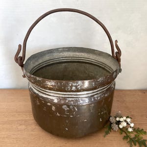 May include: An antique copper bucket with a rusty metal handle. The bucket is cylindrical with a wide rim, showing signs of age and wear. The handle is arched and attached with metal loops. A small sprig of greenery and white berries rests on the wooden surface.