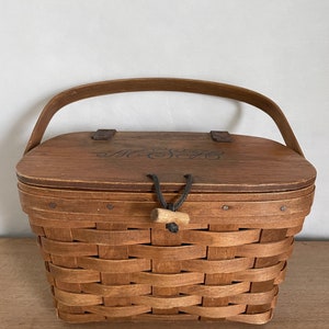 May include: A brown wooden basket with a woven pattern and a lid. The lid has a black leather handle and the words "The Sage" are engraved on the top.