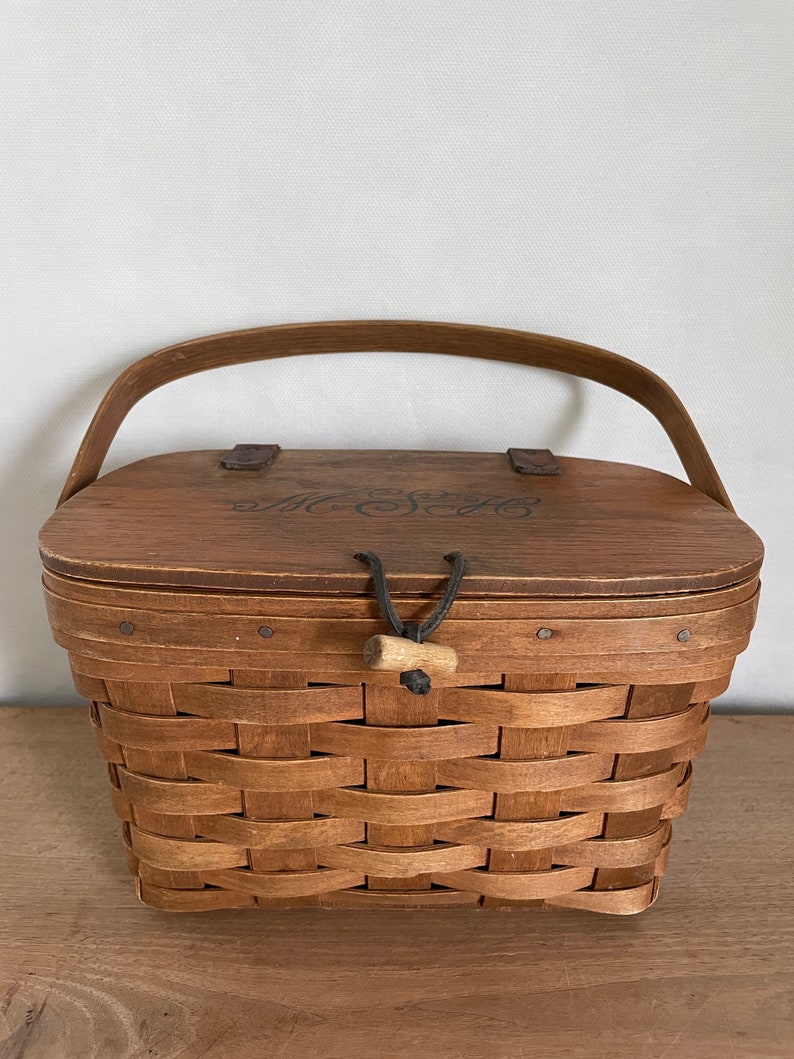 May include: A brown wooden basket with a woven pattern and a lid. The lid has a black leather handle and the words "The Sage" are engraved on the top.