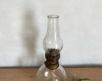 May include: A small, clear glass oil lamp with a brass wick holder and a round base. The lamp is sitting on a wooden surface with a sprig of white berries and greenery.