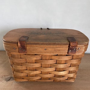 May include: A brown wooden basket with a woven pattern and a wooden lid. The lid has two leather straps with metal buckles and a floral design.