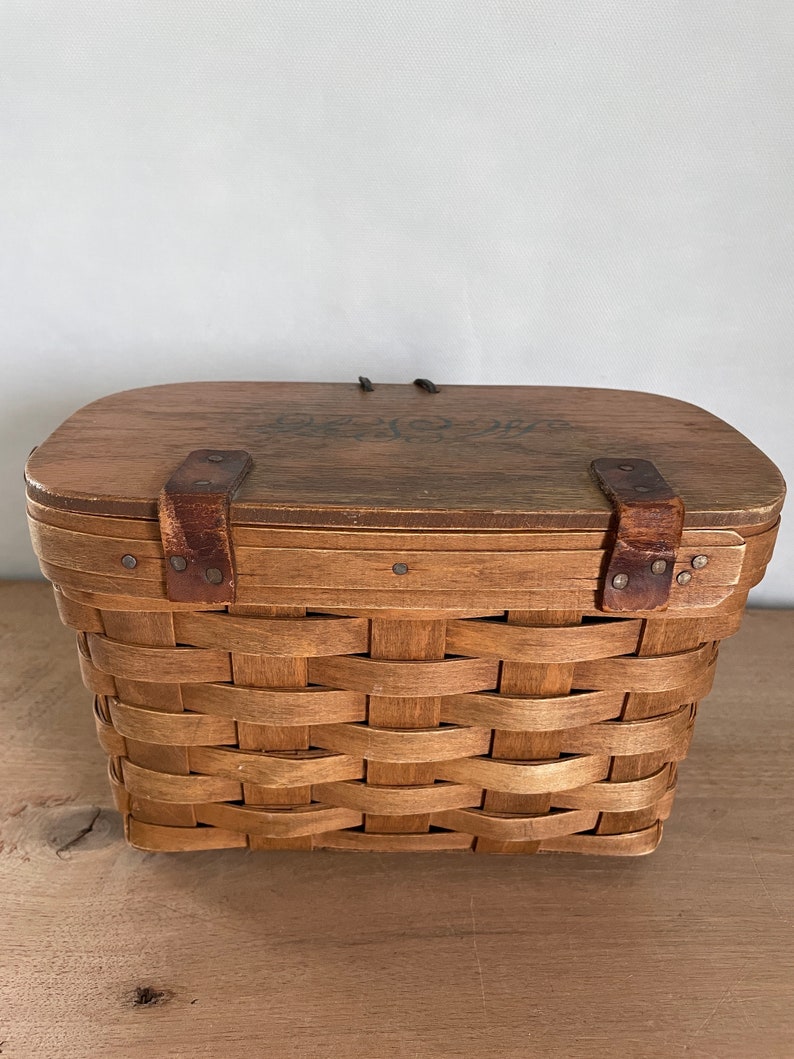 May include: A brown wooden basket with a woven pattern and a wooden lid. The lid has two leather straps with metal buckles and a floral design.