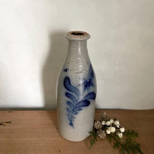 Puede incluir: Una botella de gres con un acabado gris moteado y una abertura marrón oscuro. La botella presenta un diseño floral azul y un emblema estampado cerca de la parte superior. La botella se exhibe sobre una superficie de madera.