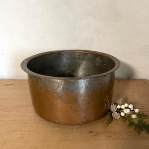 May include: A round, deep copper basin with a wide, flat rim. The exterior features a warm, metallic copper finish with visible signs of wear and patina. The interior is lined with a silver-toned metal, showing a weathered, textured surface.