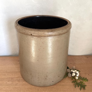 Peut inclure: Un pot en grès vintage de forme cylindrique. L'extérieur est de couleur beige moucheté, avec un intérieur marron foncé. Le bord est légèrement évasé. Une petite branche de verdure et de baies blanches est posée sur le côté.