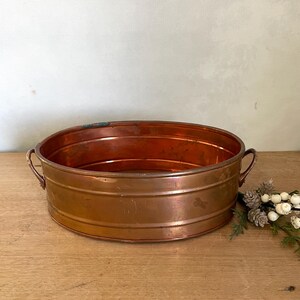 May include: A copper-colored, oval-shaped metal planter with two small handles. The planter has a ribbed design and sits on a wooden surface. Decorative sprigs of white berries and greenery are to the right of the planter.