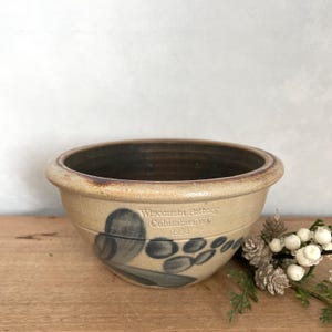 May include: A tan stoneware bowl with a dark brown interior and a blue leaf and berry design. The bowl has the text "Wisconsin Pottery Columbus, WA 1988" on the side. The bowl is on a wooden surface.