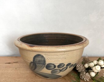 Puede incluir: Un cuenco de gres beige con interior marrón oscuro y un diseño decorativo de hojas y bayas azules. El cuenco tiene el texto "Wisconsin Pottery Columbus, WA 1988" en el lateral. El cuenco está sobre una superficie de madera.