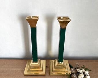 Puede incluir: Dos candelabros de latón y verde. Los candelabros tienen una base cuadrada y una parte superior cuadrada. Los tallos son verdes y las partes superiores doradas. Los candelabros están colocados sobre una superficie de madera.