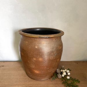 May include: A rustic, brown ceramic pot with a wide rim and a dark interior. The pot has a textured surface and sits on a wooden surface. Decorative sprigs of greenery and white berries are placed near the base of the pot.