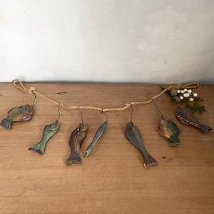 May include: A rustic garland featuring hand-painted wooden fish in various colors, including green, blue, and brown. The fish are suspended from a natural twine rope, creating a charming nautical or coastal theme. Decorative sprigs add a touch of nature.