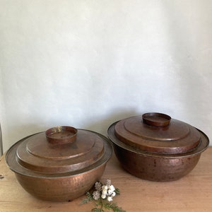 May include: Two antique copper bowls with lids. The bowls have a hammered finish and are slightly tarnished. The lids have a raised rim and a small knob on top.