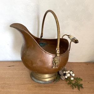 May include: Antique copper coal scuttle with a brass base and handle. The scuttle features a spout, a decorative lion's head, and a ceramic handle with blue and white floral patterns. A vintage home decor item.