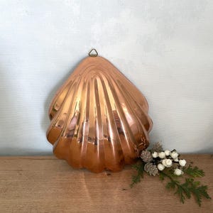 May include: A polished copper mold shaped like a large seashell with vertical ridges and a small metal hanging loop at the top. The metallic surface reflects light, showing a warm, reddish-brown tone throughout the curved, fan-like structure.