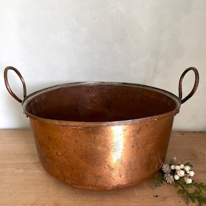 May include: A large, antique copper bowl with two curved handles. The bowl has a wide, open design and a rich, reddish-brown color. The rim is slightly darker, and the interior shows signs of age. A decorative sprig of white berries and greenery sits on the wooden surface.