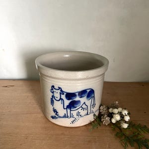 May include: A cylindrical ceramic crock with a light beige finish and a blue hand-painted cow illustration on the front. The vessel features a smooth interior, a slightly flared rim, and a sturdy base, providing a functional container for storage.
