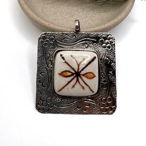 May include: A square pendant with a silver-colored frame and a cream-colored center. The center features a brown and orange design. The pendant is displayed on a small, round, beige dish with a green pine needle sprig.