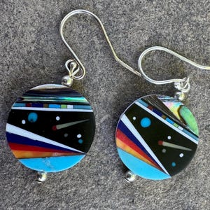 May include: A pair of round dangle earrings featuring silver-tone hooks and circular pendants. The pendants display a geometric inlay pattern with black, turquoise, red, orange, and white segments, accented by small dots and thin lines.