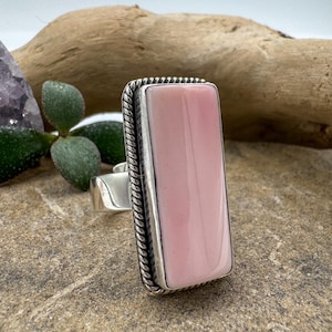 May include: A rectangular pink gemstone ring with a silver band. The gemstone is a light pink color and is set in a silver bezel. The ring has a twisted rope design around the gemstone. The ring is on a stone surface.